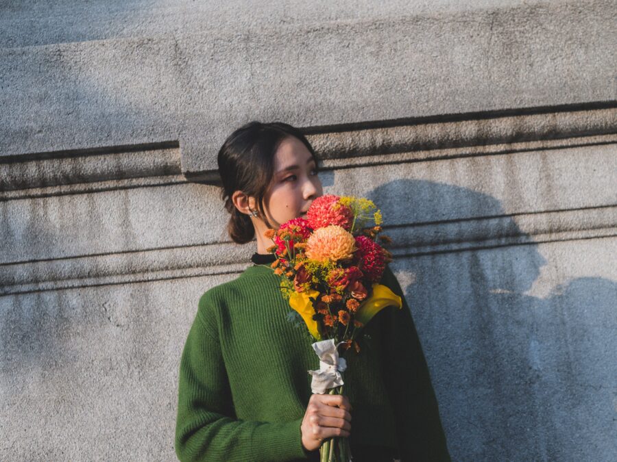 穿綠色毛衣的女子捧著花束，影子落在石牆上